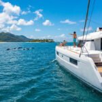 Catamaran à Maurice avec passagers regardant des dauphins, femme avec masque de plongée, plages tropicales et montagnes en fond.