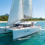 Grand catamaran blanc avec des passagers naviguant sur des eaux turquoises près d'une plage tropicale et de montagnes verdoyantes.