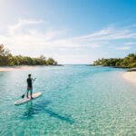 Un homme fait du paddle sur des eaux turquoises et peu profondes, longeant une plage de sable blanc bordée de palmiers sous un ciel ensoleillé.